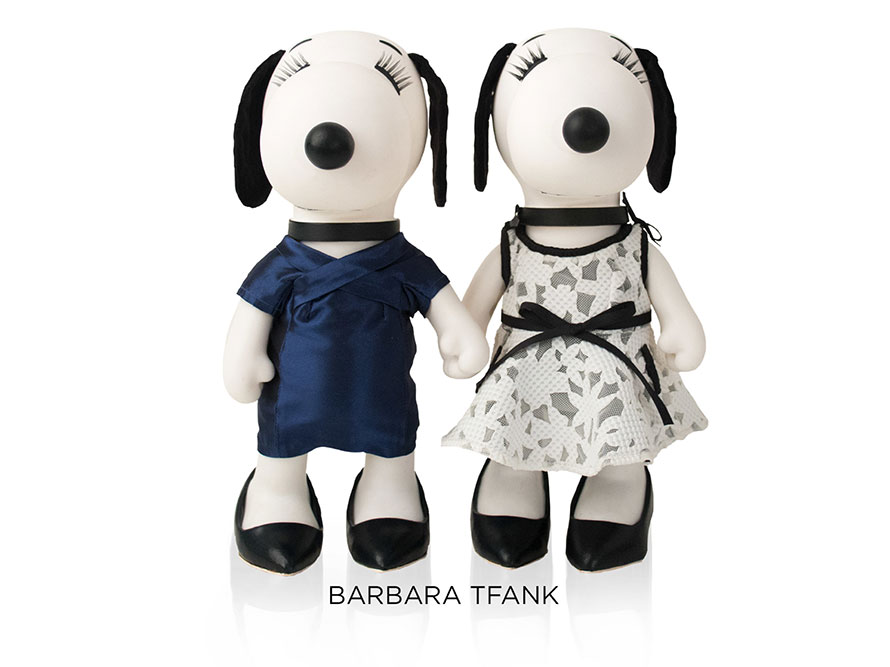 Two black and white, stuffed toy dogs standing and holding hands in front of a white background. The dog on the left is wearing a dark blue dress and the dog on the right is wearing a white dress with black detailing.