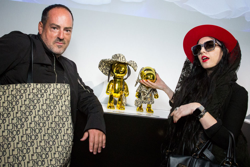 A man and woman posing for a photo at an event. The man on the left is wearing a black jacket and a big purse. The girl on the left is wearing a red hat, black clothes and touching a gold statues beside her.
