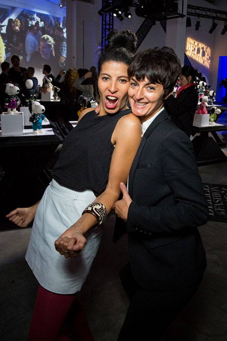 Two women posing for a photo at a semi formal event. There are people behind them, a large screen projection and small black and white dog statues on display.