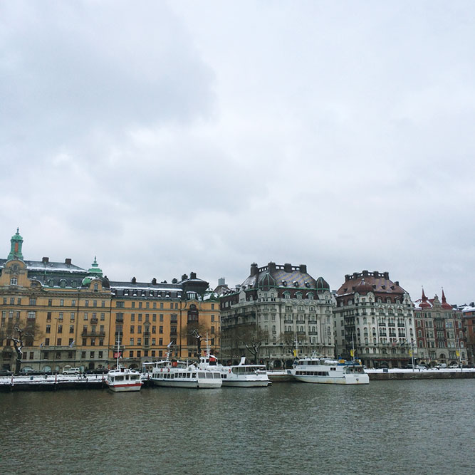 A waterfront, European city on a cloudy day with several boats docked at the pier.