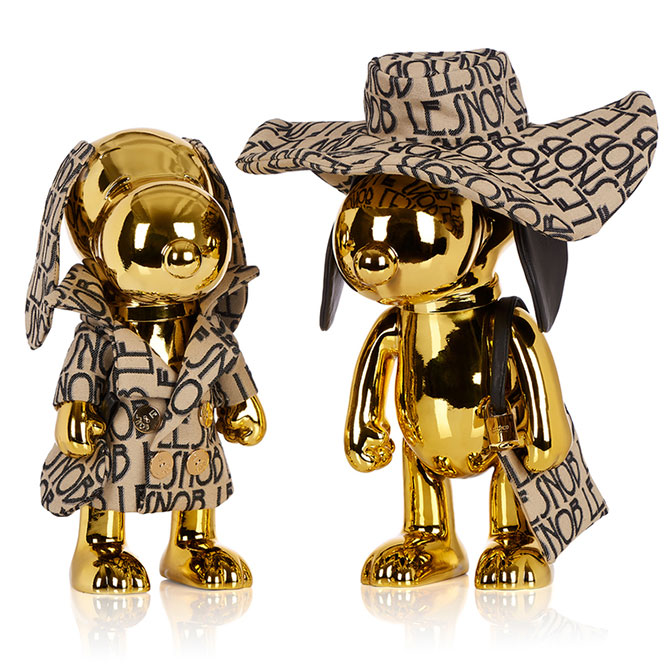 Two gold dog statues in front of a white background wearing matching beige and black outfits.