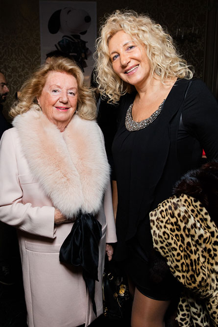 Two blonde, older woman posing for a picture at an event. The women on the left is wearing a light pink coat and the woman on the right is wearing a black dress.