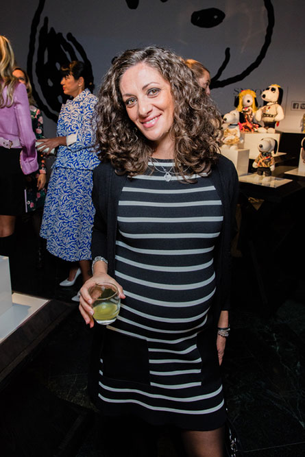 A woman wearing a black and white striped dress, with curly brown hair, is holding a drink in her hand and posing for a photo.