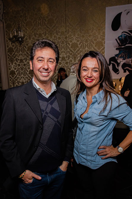 A brunetter woman wearing a denim shirt is standing beside a man wearing a black jacket. They are posing for a photo at an event.