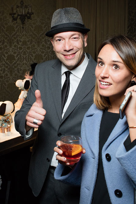 A man and a woman at a semi-formal event. The man is wearing a grey suit, a hat and giving a thumbs up. The girl is wearing a blue jacket and holding a cocktail in her hand.