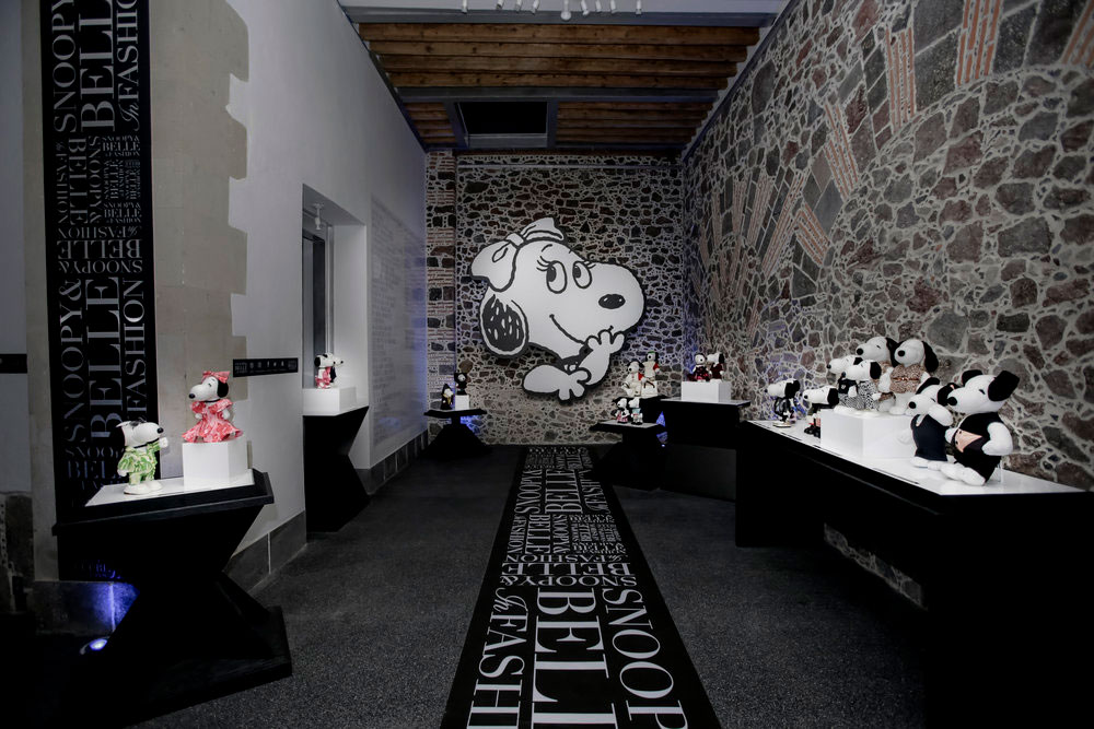 An indoor space with brick walls, grey carpets and black tables displaying black and white dog statues wearing various costumes. At the very back, there is a large art piece on the wall of a black and white dog.