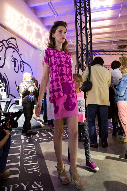 A tall, lean woman modelling a short pink dress at an indoor event space. There are people in the background.