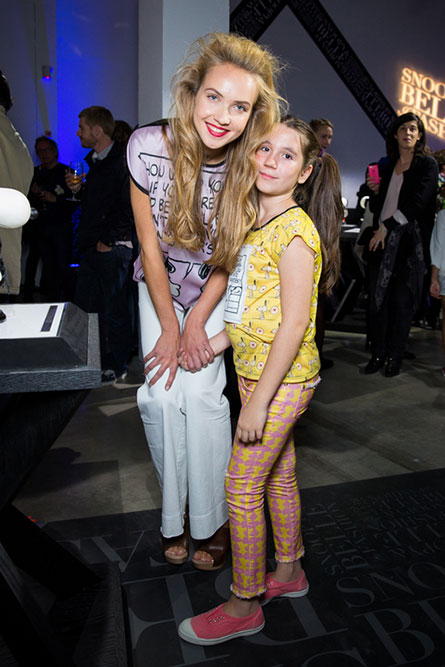 A tall, blonde woman is leaning over and standing beside a young girl. They are posing for a photo at an event.