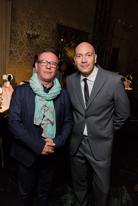 Two men standing beside each other posing for a photo at an event. The man on the right is wearing a grey suit and the man on the left is wearing a light blue scarf and dark blue jacket.