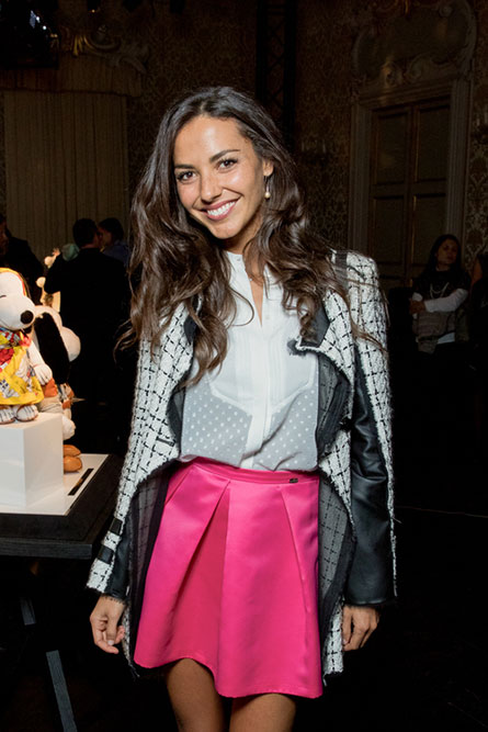 A tall, brunette woman posing for a photo at an event. She is wearing a white blouse, a pink skirt and a grey jacket.