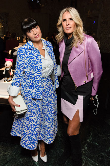 A brunette woman, wearing a blue and white dress is standing beside a blonde woman wearing a black dress with a pink jacket. They are posing for a picture at an event.