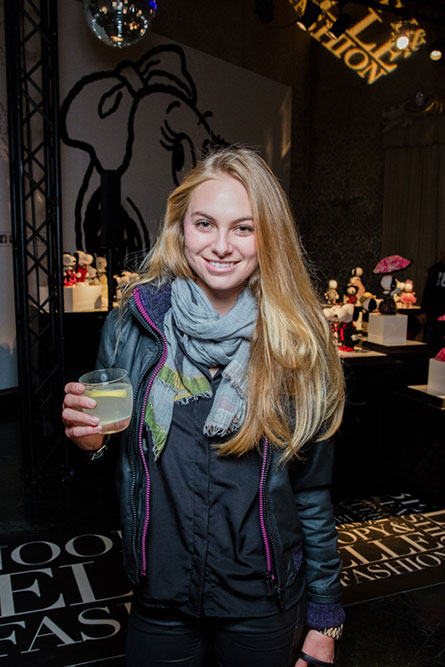 A woman with long blonde hair, wearing a black jacket and a grey scarf, posing for a photo and holding a cocktail in her hand.