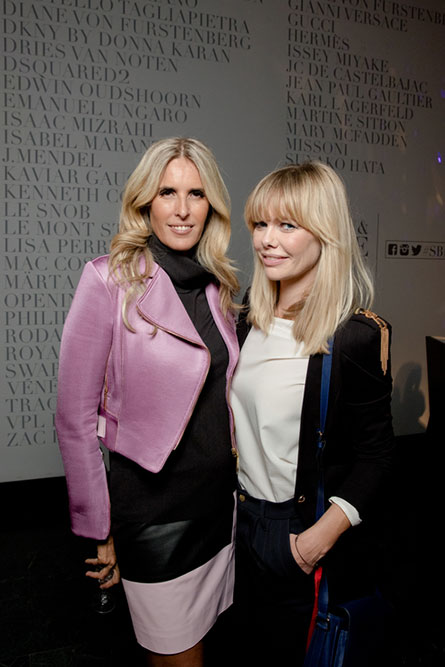 Two middle-aged, blonde women posing for a photo at an event. The woman on the left is wearing a pink jacket and the woman on the right is wearing a white blouse and a black jacket.