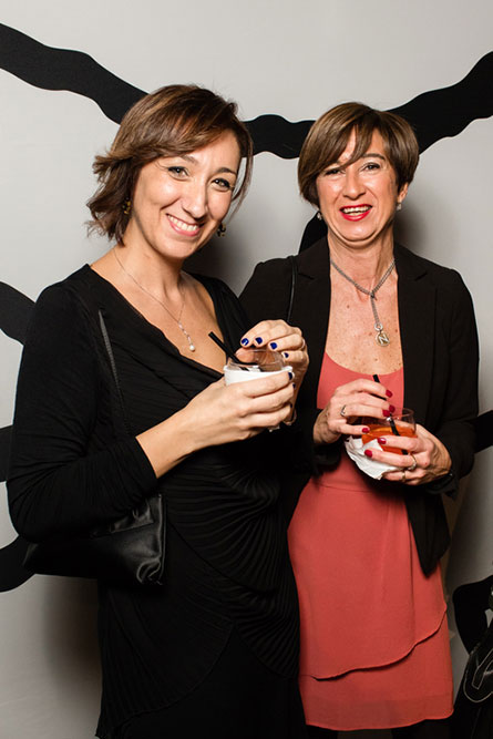 Two middle aged women, wearing semi-formal attire, are holding cocktails in their hands and posing for a photo at an event.
