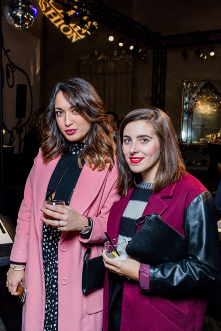 Two women posing for a photo at an event and holding cocktails in their hands. The woman on the left is wearing a light pink coat and the woman on the right is wearing a dark pink coat.